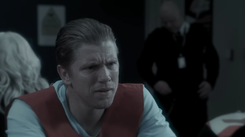 A man with light hair wearing an orange vest over a light blue shirt looking distressed in a dimly lit room.