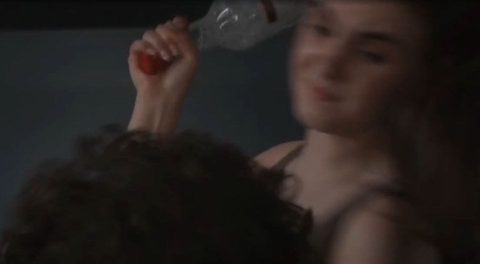 A woman holds an empty glass bottle with an orange lid above another person's head, ready to strike.