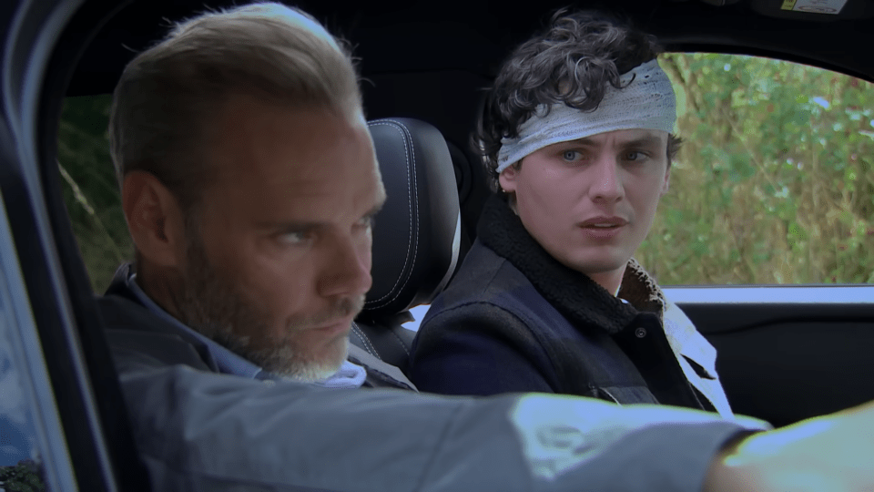 Two men in a car, the driver looking focused, and the passenger with a bandage on his head looking forward with wide eyes.