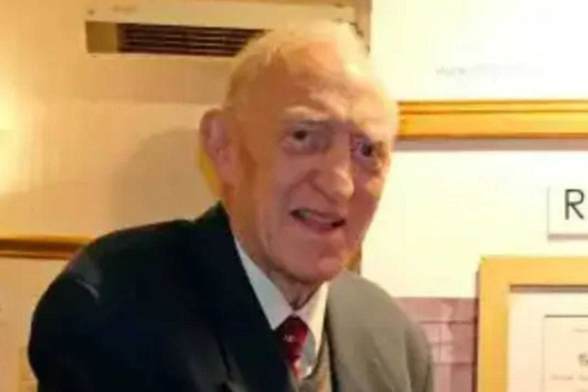 An image collage containing 1 images, Image 1 shows A man in a suit jacket and tie looks at the camera, with framed documents behind him
