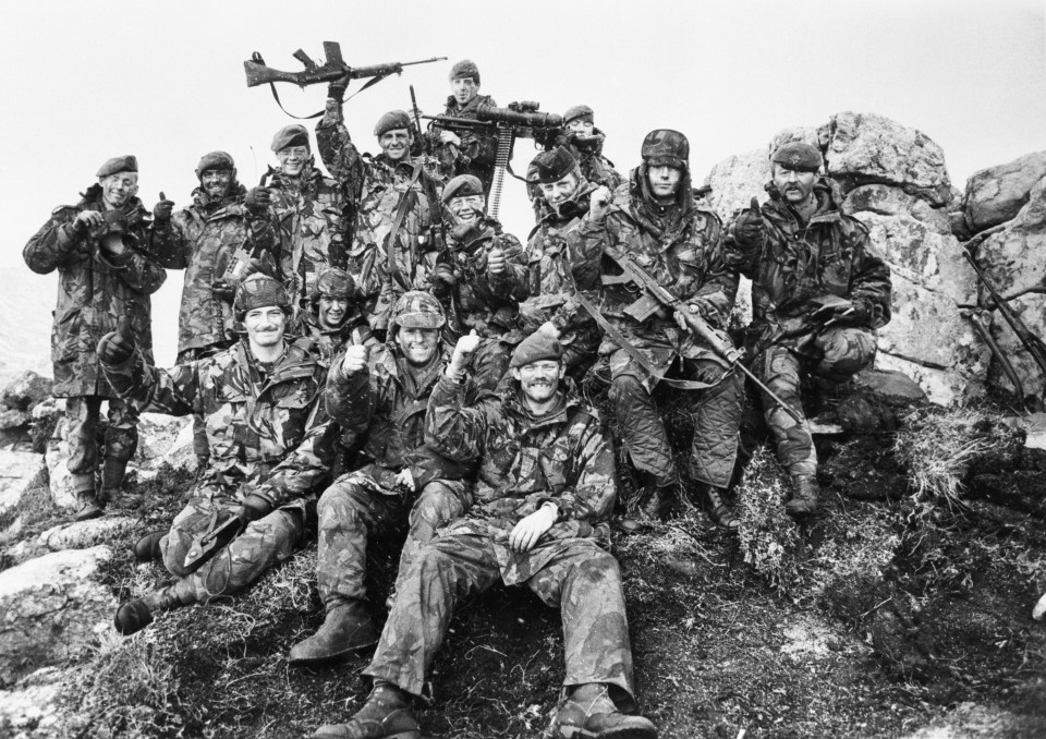Black and white photo of a platoon of soldiers, some with weapons, giving thumbs up.