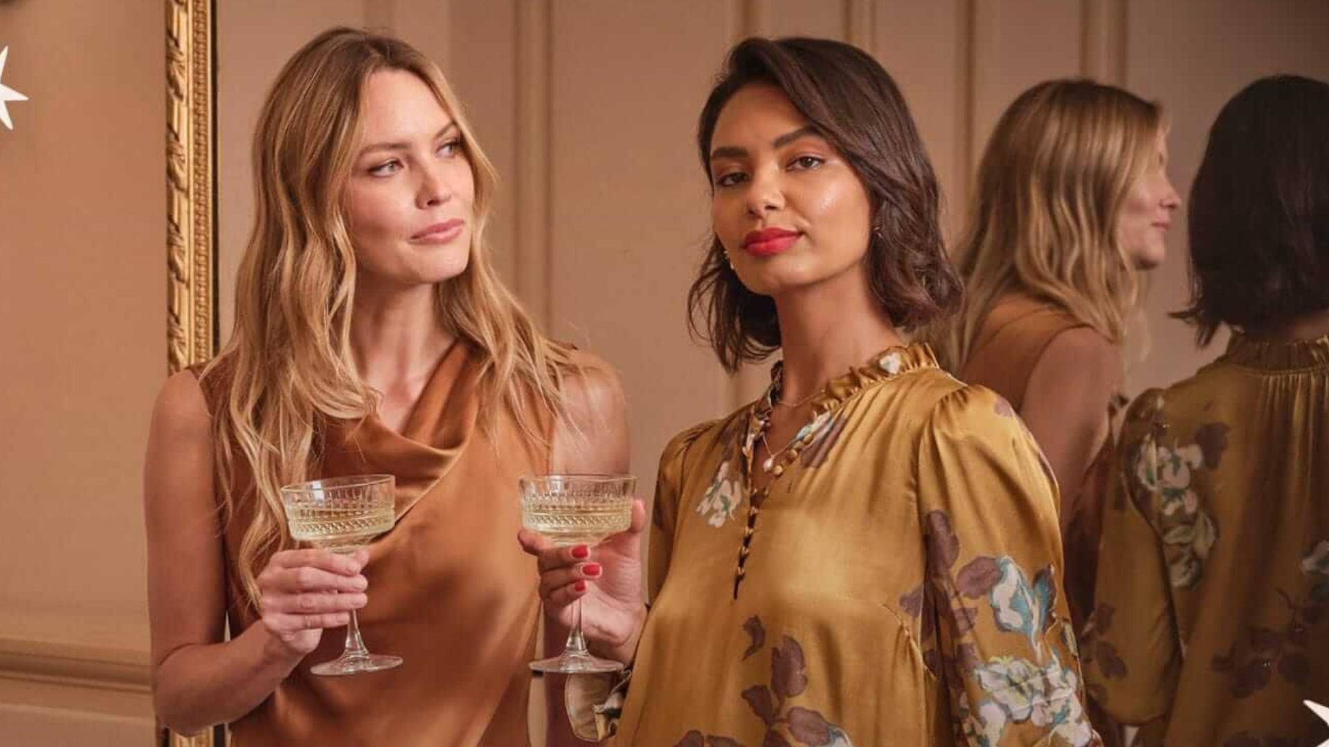 Two women holding champagne glasses, with one woman looking directly at the camera and the other looking away, and a third woman's reflection in a mirror.