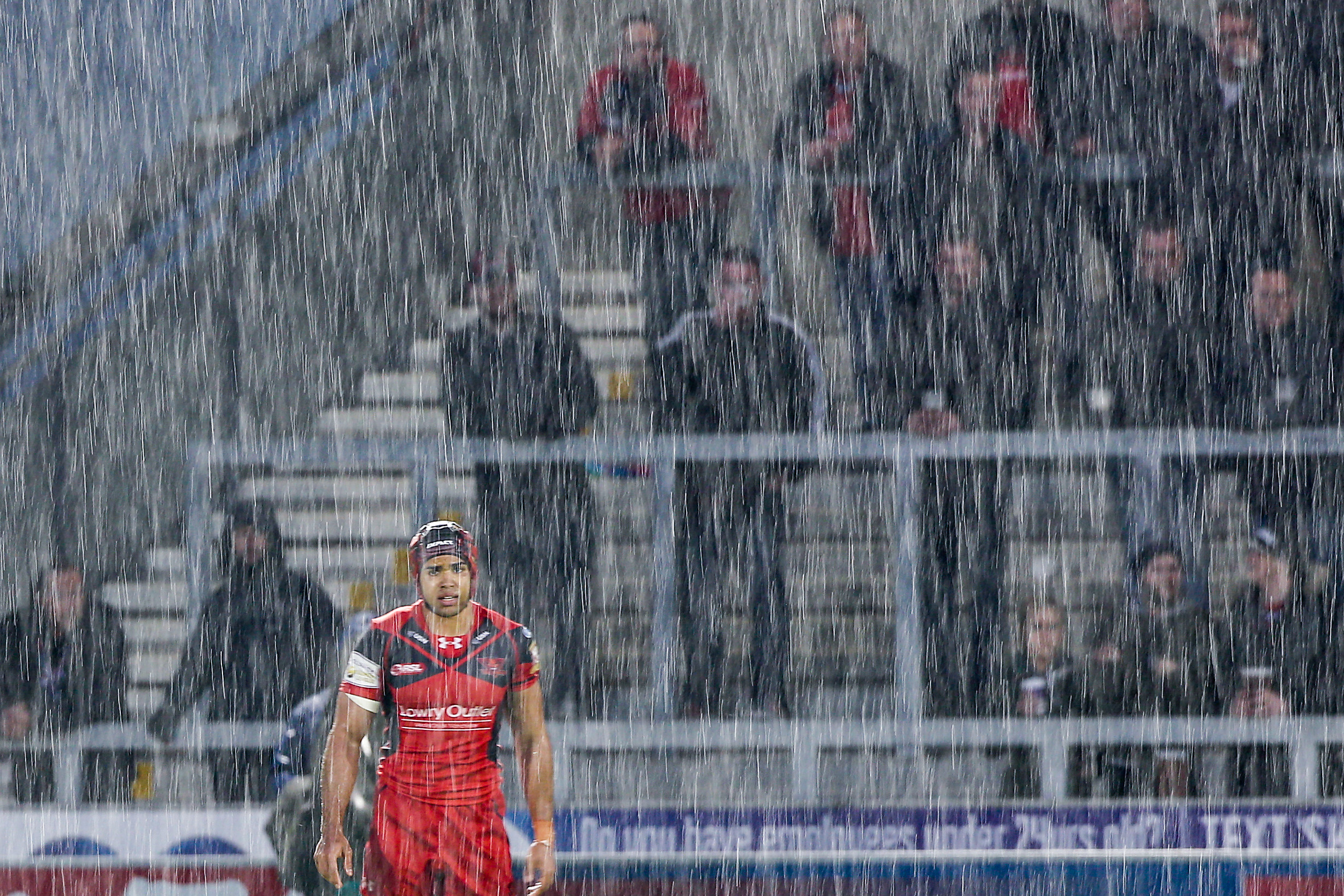 Salford Red Devils player in red and black uniform standing on a rainy field with blurred fans in the stands behind him.