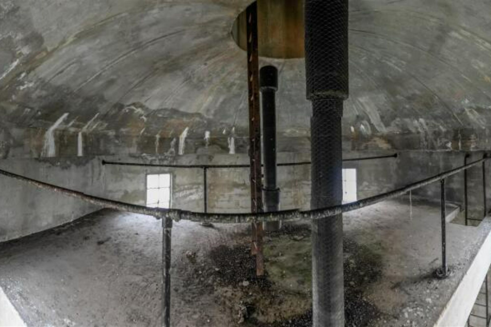 An image collage containing 1 images, Image 1 shows Interior of an old water tower with concrete walls and ceiling, two large pipes in the center, and small windows