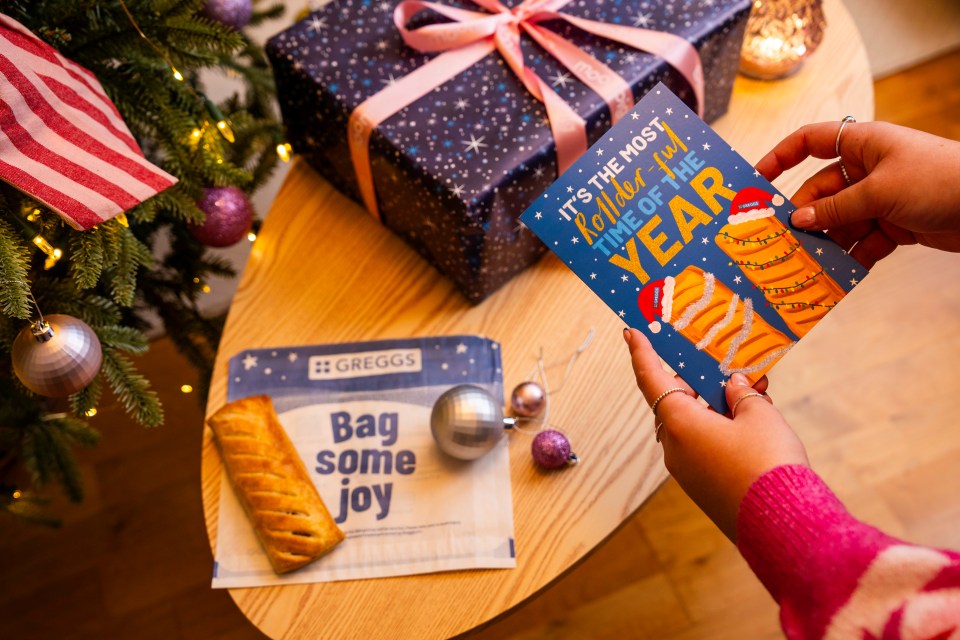 A hand holds a Greggs Christmas card that says "IT'S THE MOST Roller-ful TIME OF THE YEAR" with two sausage rolls wearing Santa hats.