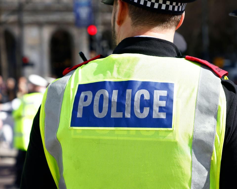 Back of a police officer wearing a neon yellow reflective vest over a dark uniform with "POLICE" written in reflective letters on a blue rectangle.