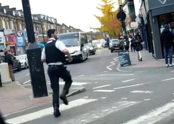 A police officer appears to run straight past on-the-run Algerian sex offender Brahim Kaddour-Cherif who can be seen standing on the corner of the street in a grey hoodie