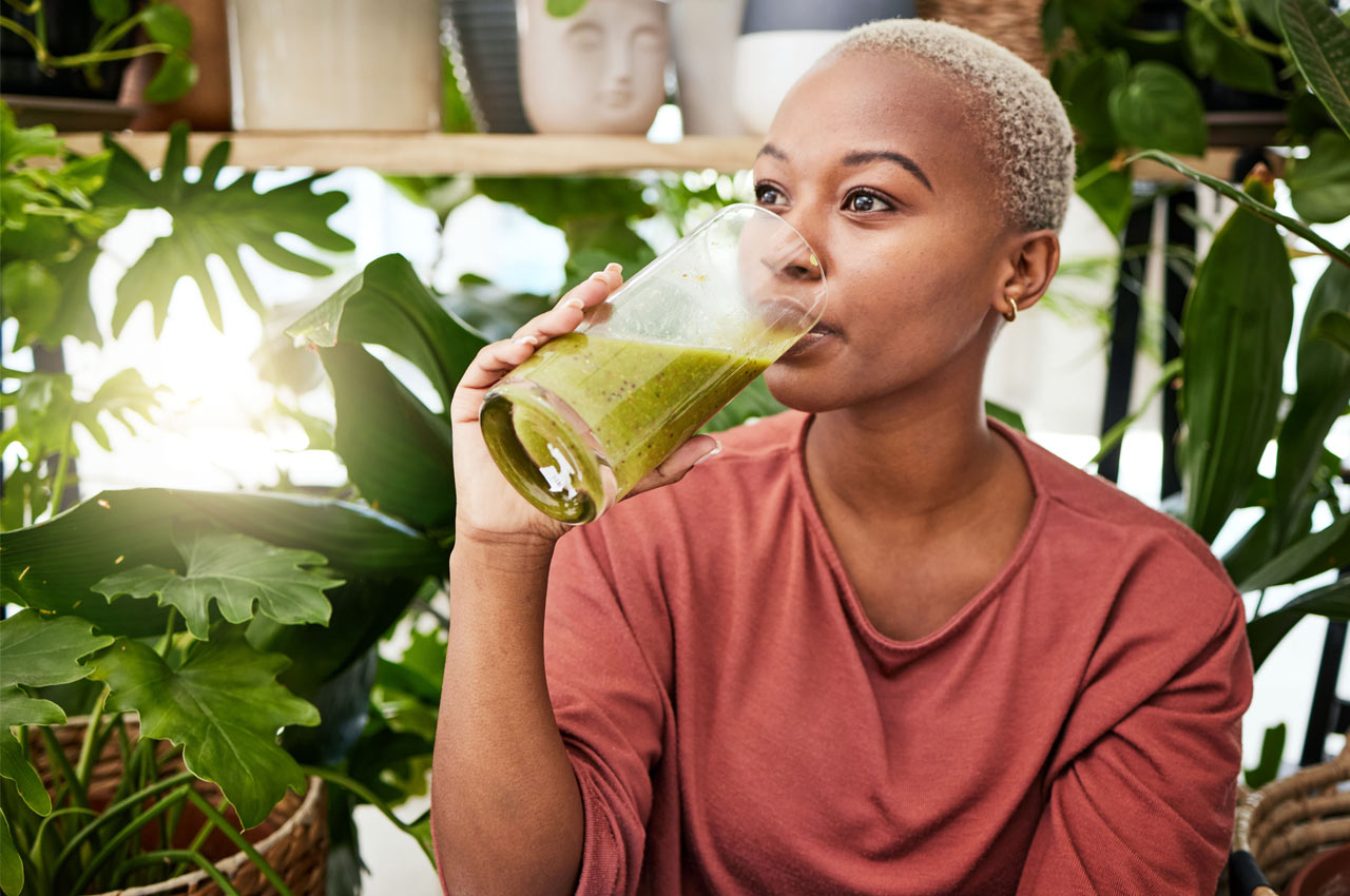 Woman drinking a green smoothie surrounded by plants.