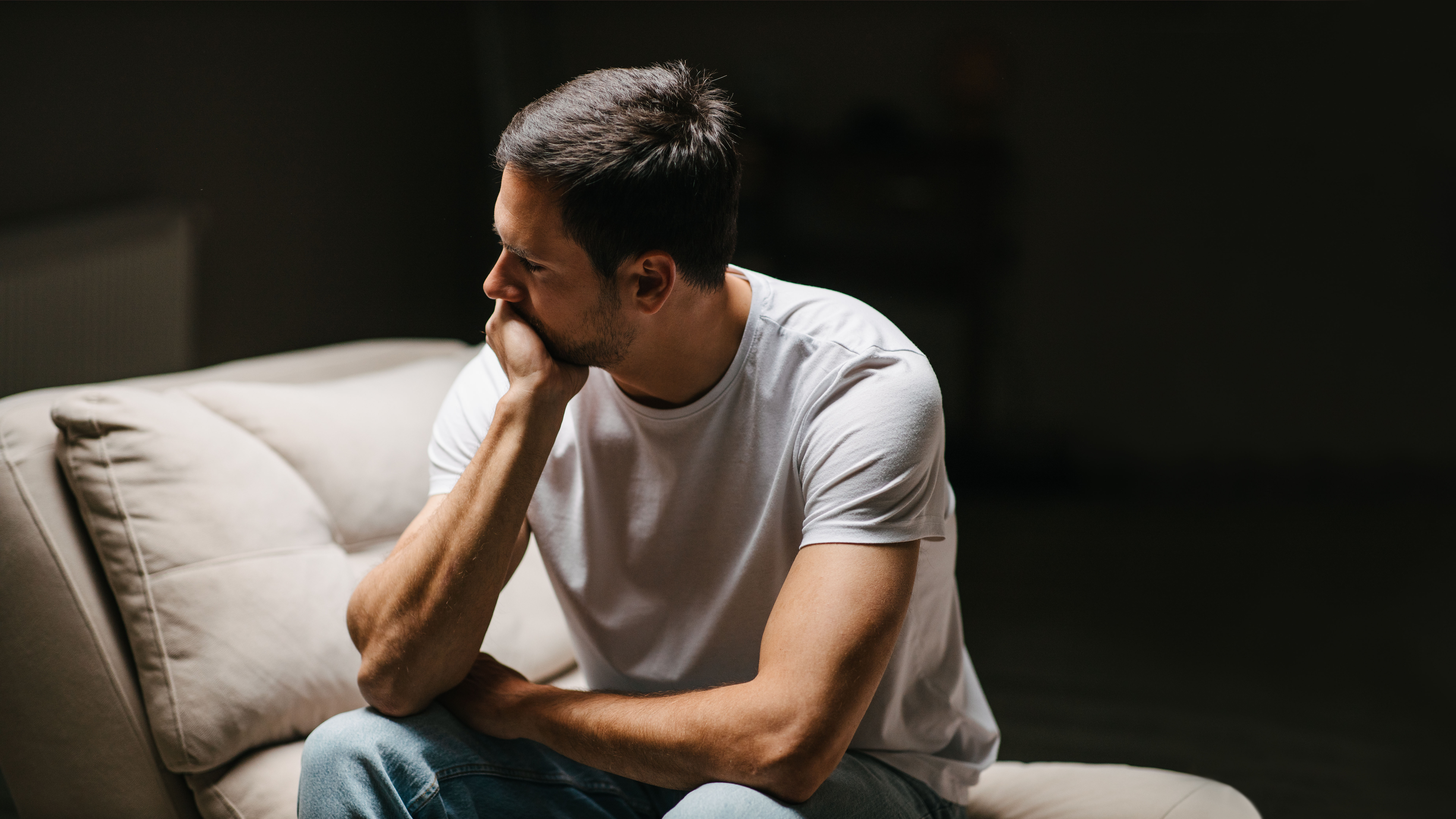 Depressed young man lost in his thoughts, sitting on a sofa.