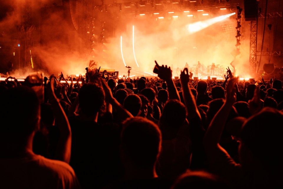 A crowd of people with raised hands at a concert with an orange-lit stage and smoke.
