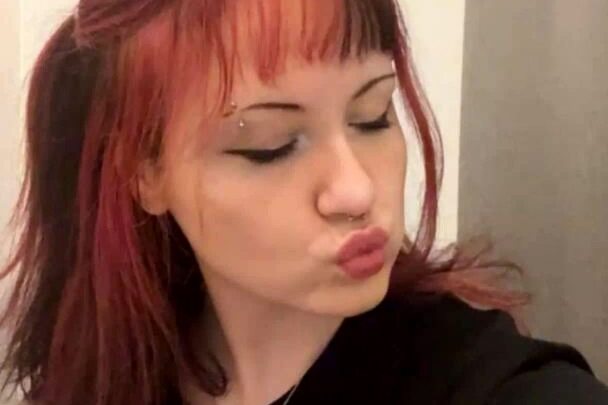 An image collage containing 1 images, Image 1 shows A person with red and black hair, eyebrow and nose piercings, and dramatic eyeliner, wearing a black t-shirt with a cartoon character, poses while text overlaid on the image states, "currently thinking about the time i got such a bad tattoo that the artist paid me not to post it anywhere."