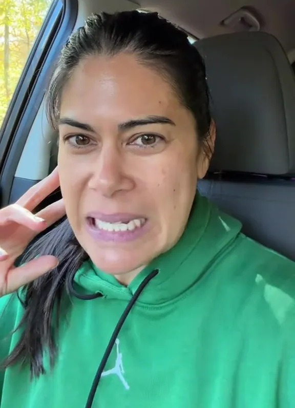 A woman in a green hoodie grimacing in a car.