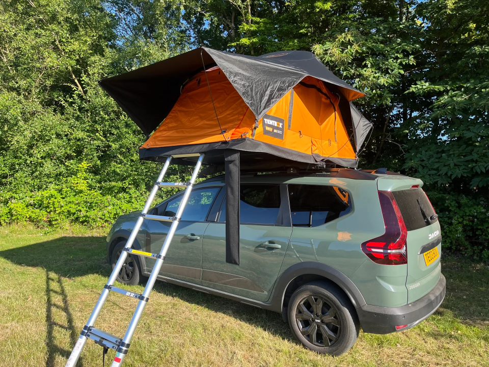A green Dacia Jogger with an orange and black rooftop tent set up.