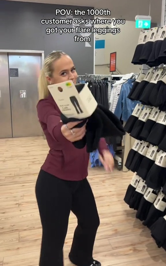 A blonde woman in a burgundy top and black flare leggings holds up a packaged pair of black flare leggings in a store, with the text "POV: the 1000th customer asks where you got your flare leggings from" overlaid.