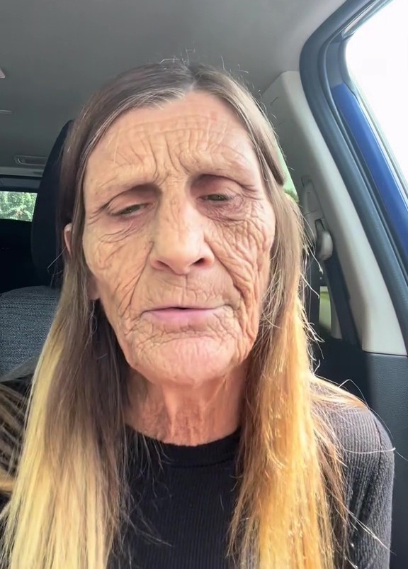 A woman with long light brown hair wears an aging filter over her face, sitting in a car.