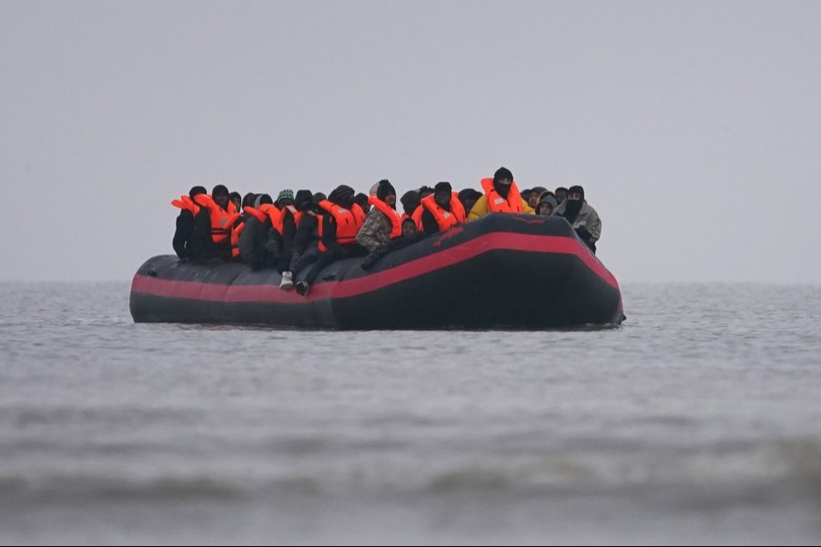 A black and red inflatable boat filled with people wearing orange life vests on the water under a cloudy sky.