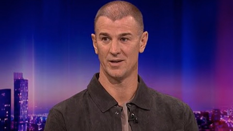 Man with a shaved head and a dark collared shirt speaking against a city skyline backdrop.