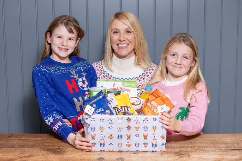 Lynsey Hope and her children, Olive and Ivy, smile as they hold a box filled with items for a reverse advent calendar, including M&M's, "Harvesst Morn" cereal, and chocolate spread.