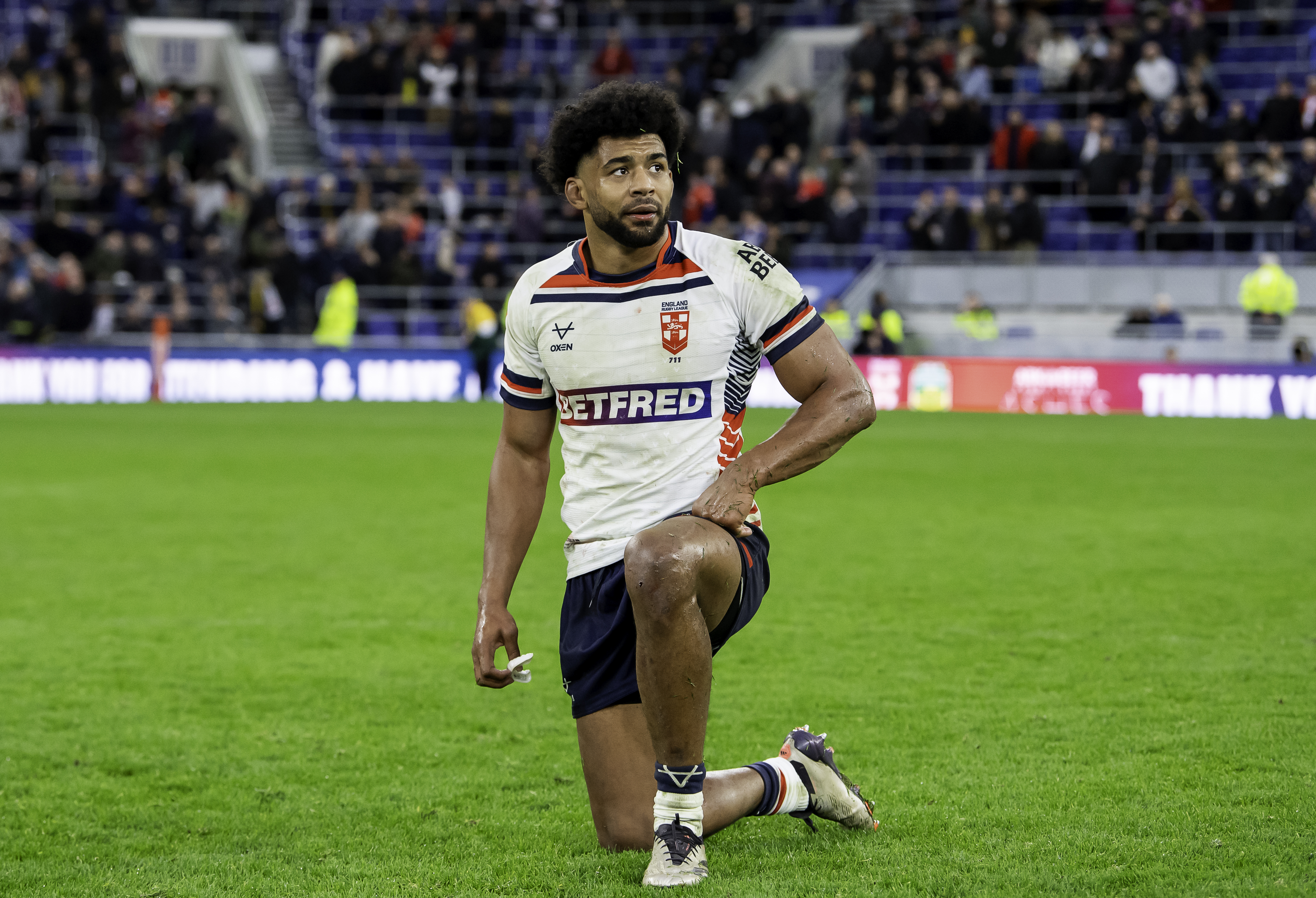 A male rugby player in a white jersey with England Rugby League emblem, kneeling on the grass pitch and looking up.
