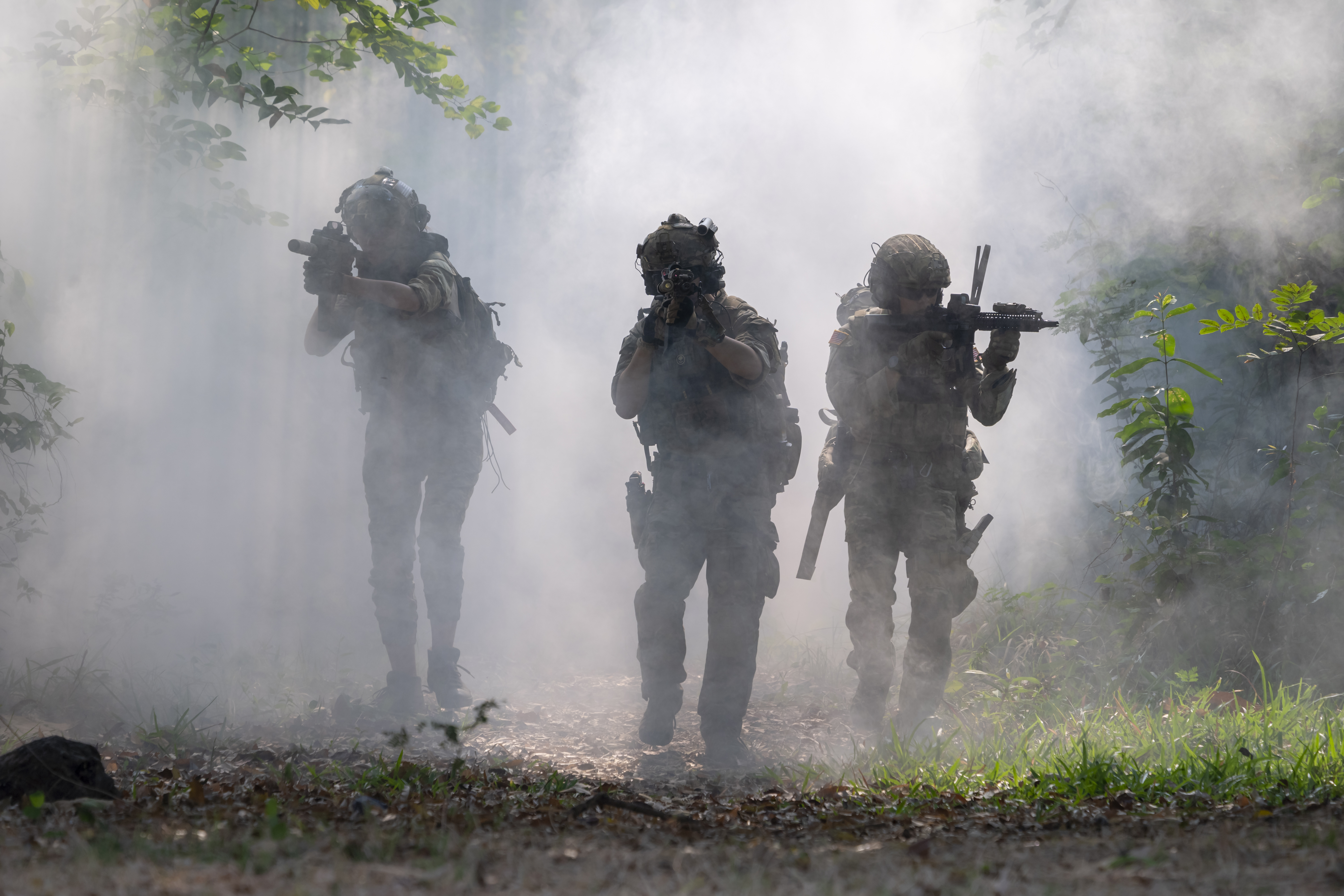 Three armed soldiers in military uniforms walking through heavy smoke in a forest.