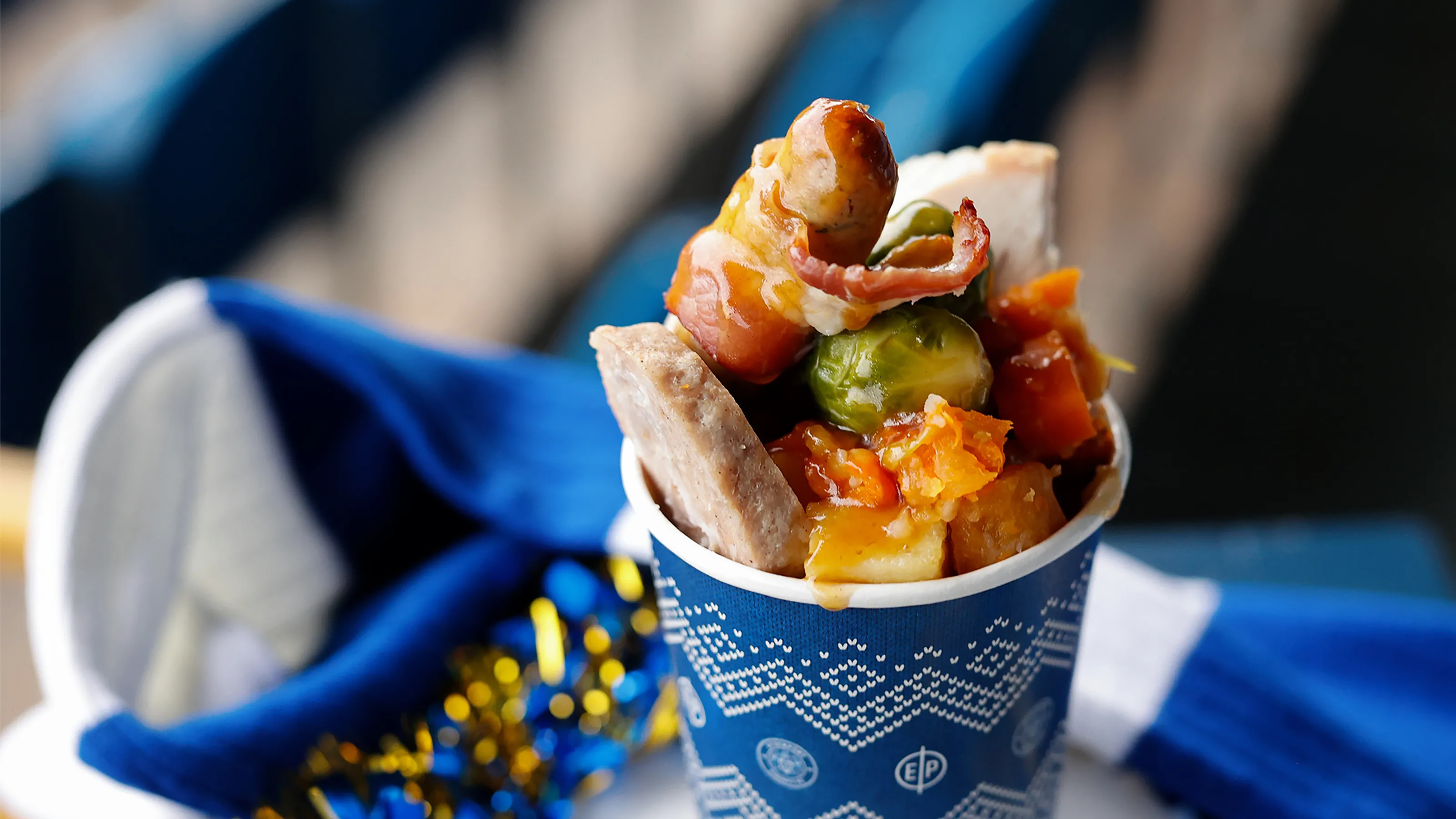 A close-up of a blue paper cup filled with Christmas dinner elements, including a pig in a blanket, Brussels sprouts, and gravy, set against a blurred background of blue and white fabric.