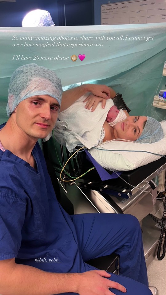 A man in blue scrubs and surgical cap takes a selfie with a woman in a hospital bed, also in a surgical cap, holding a newborn baby.