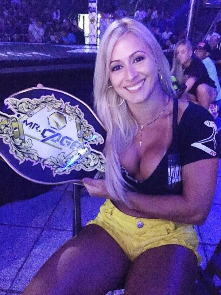A woman with blonde hair in a black top and yellow shorts holding a "MR. CAGE" championship belt, smiling for a photo.