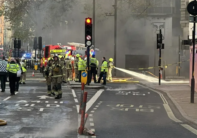 Firefighters working at a fire scene with smoke and emergency vehicles visible.