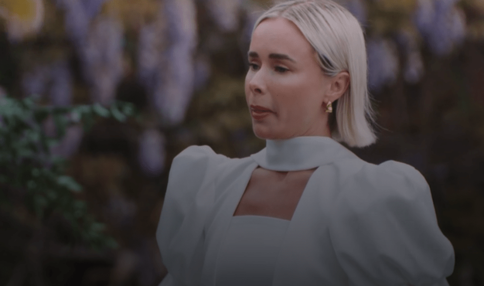 A fair-skinned woman with short blonde hair and a white dress looks off-camera.
