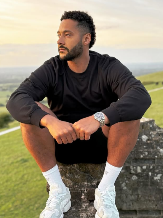 A man in a black sweatshirt, black shorts, and white sneakers sits on a stone structure outdoors, looking to his right.