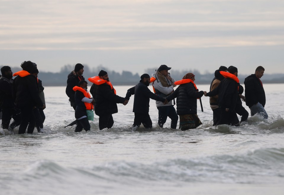 Migrants wearing orange life jackets wading through the shallow water of a sea.