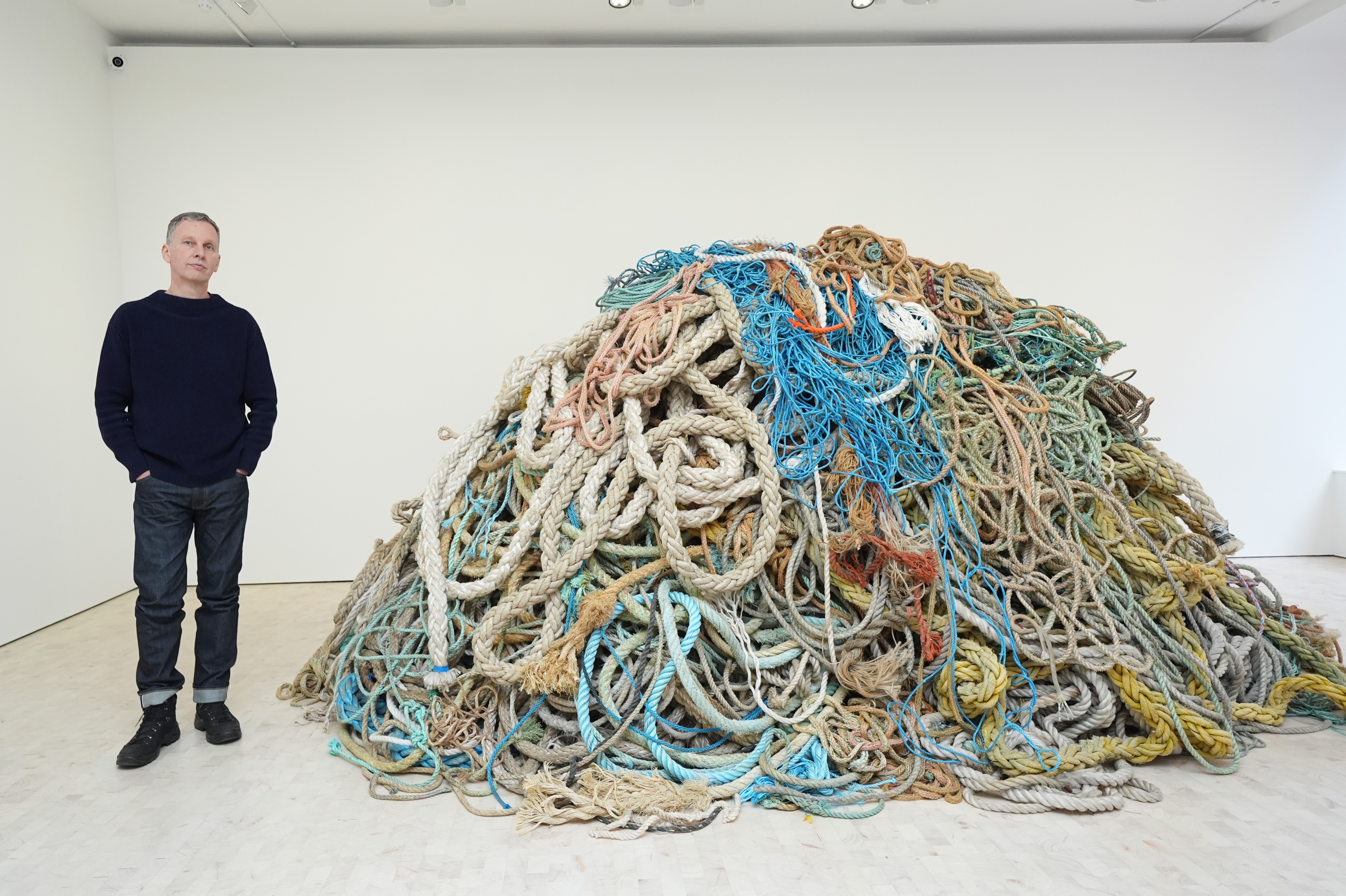 Artist David Shrigley stands beside his artwork, a large pile of multicolored ropes.