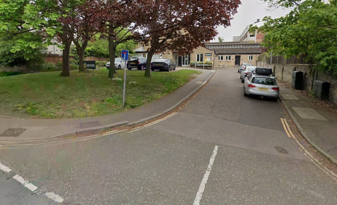 A two-lane road curves left, with parked cars on the right and a grassy area with trees and a building in the background on the left.