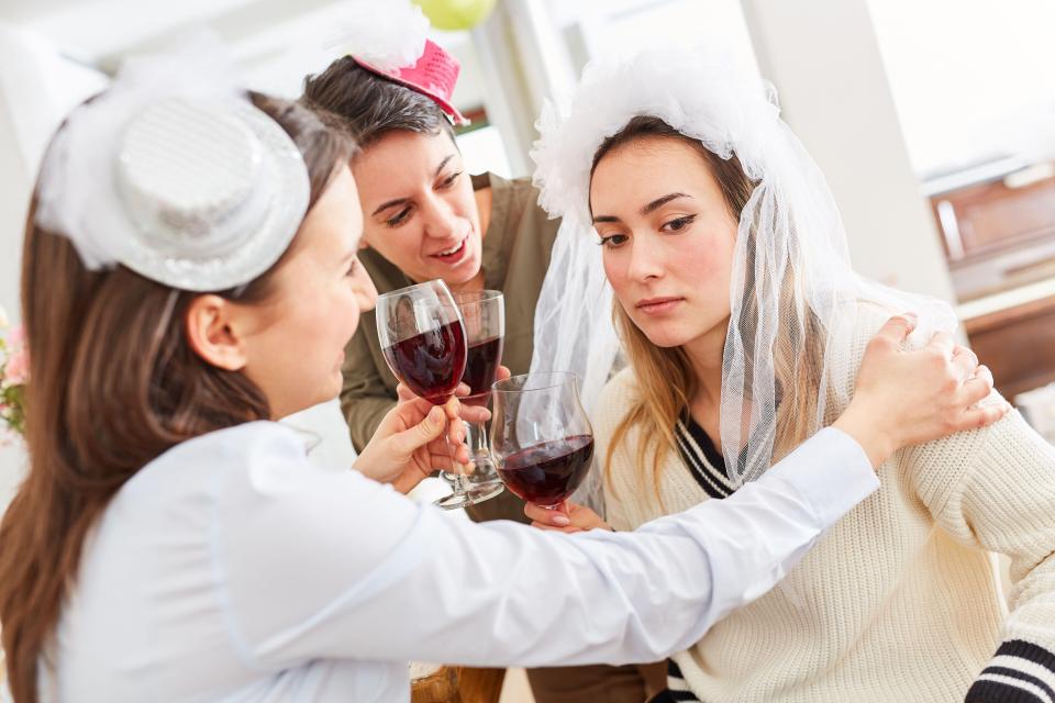 Two friends consoling a sad bride at a bachelorette party with red wine.