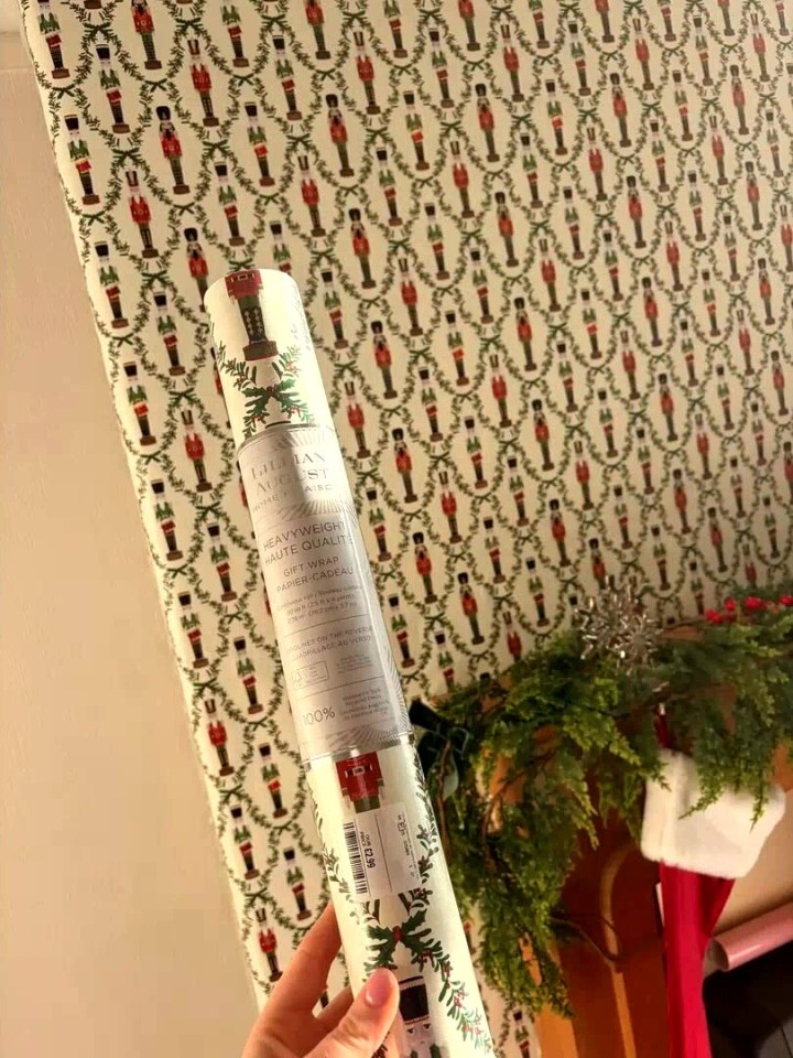 A hand holds a roll of gift wrap patterned with nutcrackers and holly, matching the wallpaper behind it.