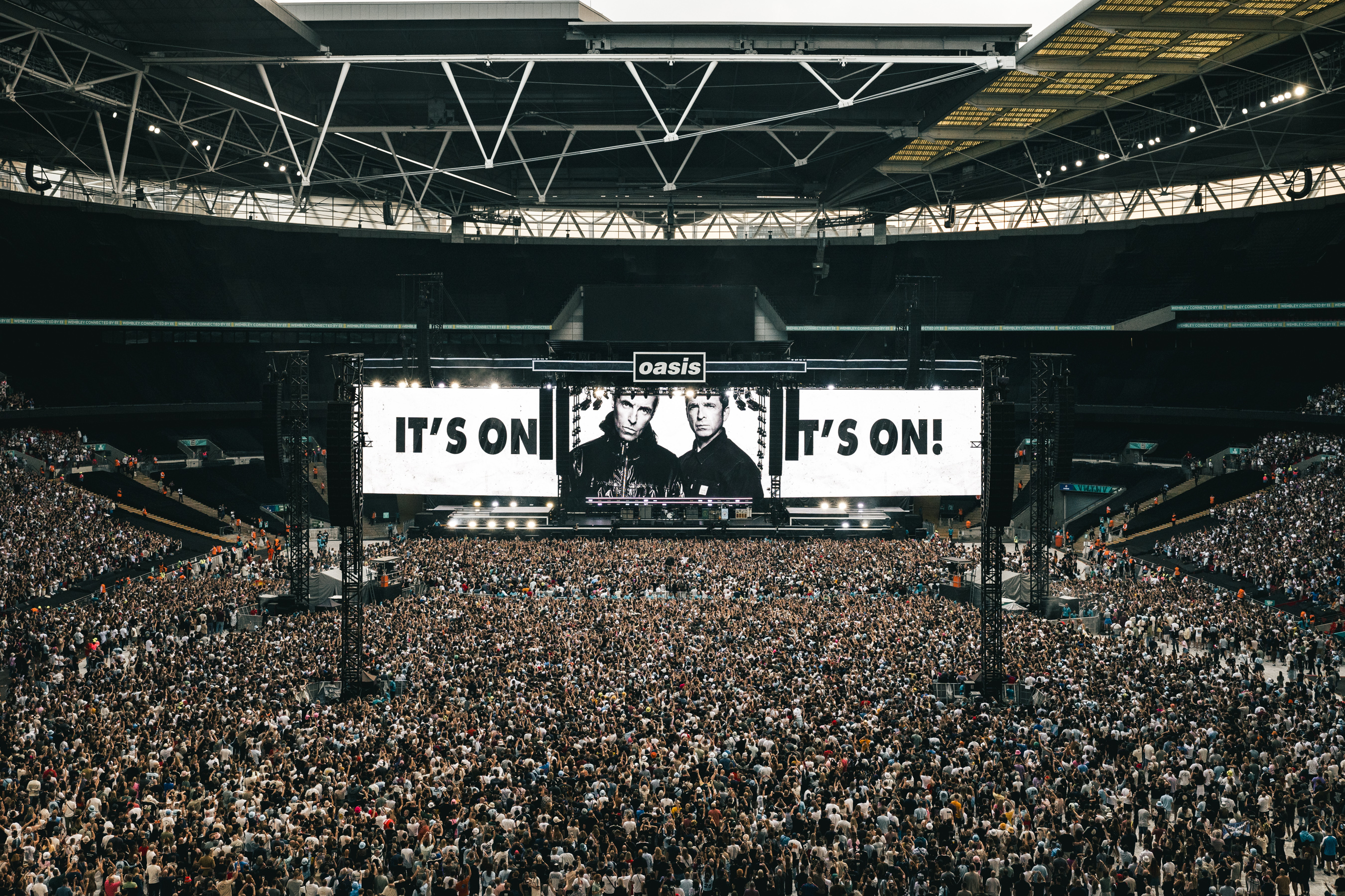 A stadium filled with a massive crowd watching a large screen displaying "Oasis It's On!" and a picture of Liam and Noel Gallagher.