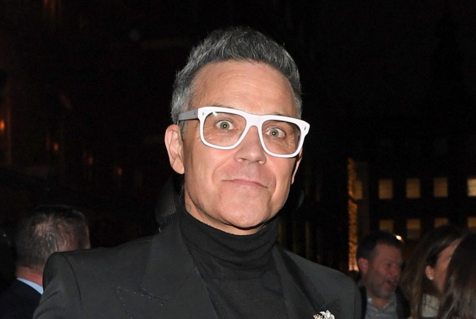 Robbie Williams in a black suit with a black turtleneck and white glasses, reaching out his hand.