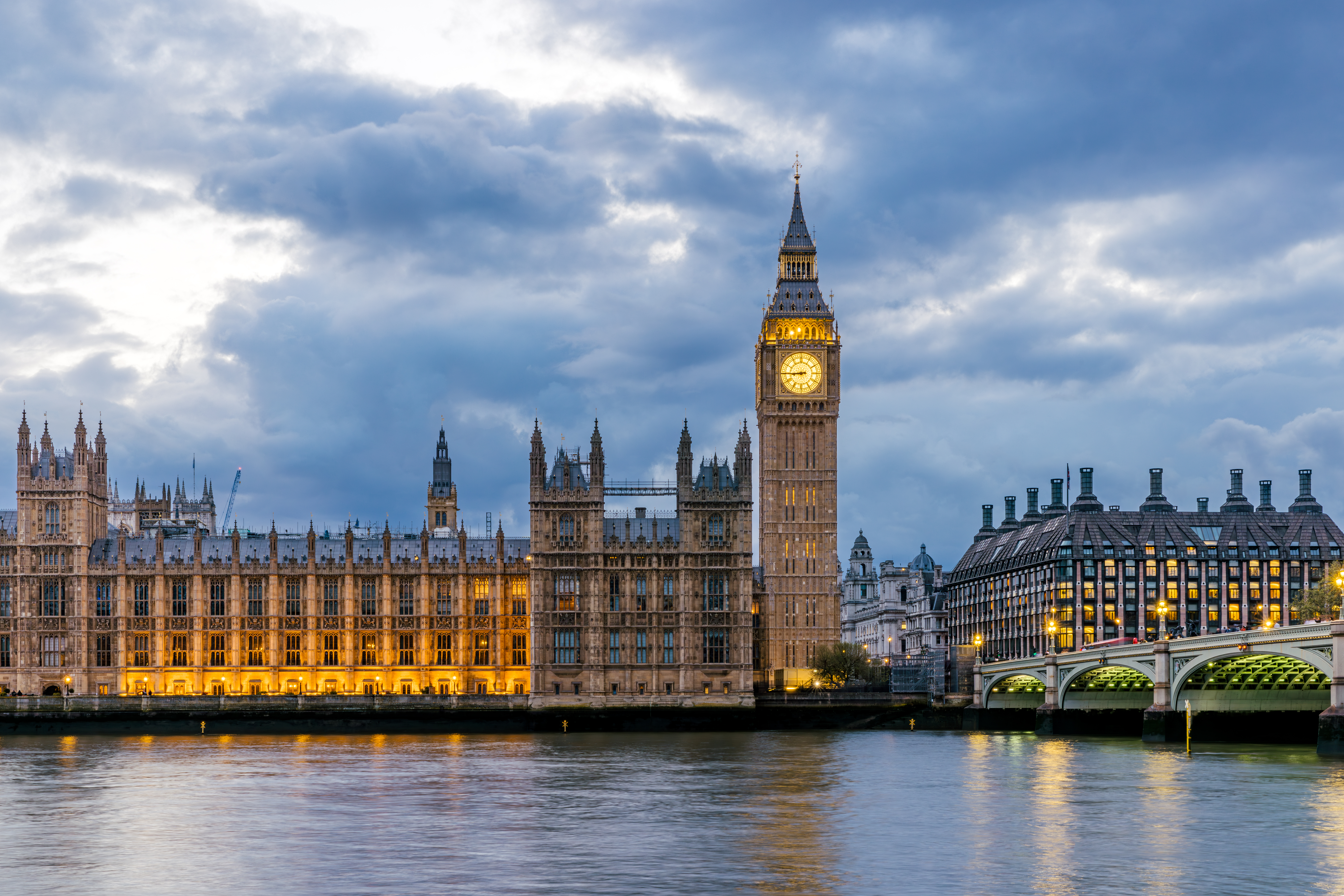 Illuminated Big Ben and the Houses of Parliament at twilight with the Thames River in the foreground.