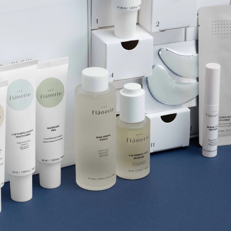An assortment of Flânerie skincare products, including tubes, bottles, and under-eye masks, on a blue surface with an advent calendar in the background.