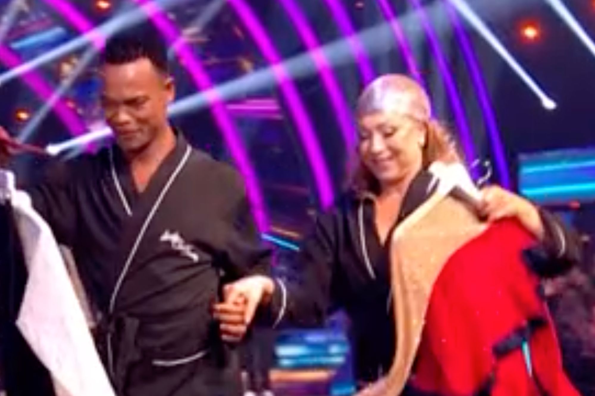 An image collage containing 1 images, Image 1 shows Two dancers in bathrobes walk across a stage carrying costume dresses, with racks of costumes behind them