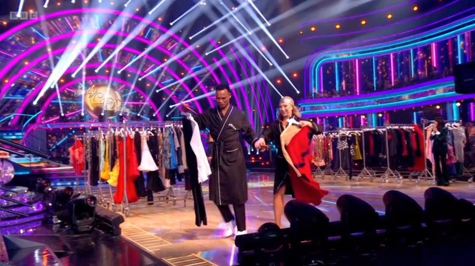 Two people wearing black robes on a dance floor, holding clothes, with clothing racks and bright lights in the background.