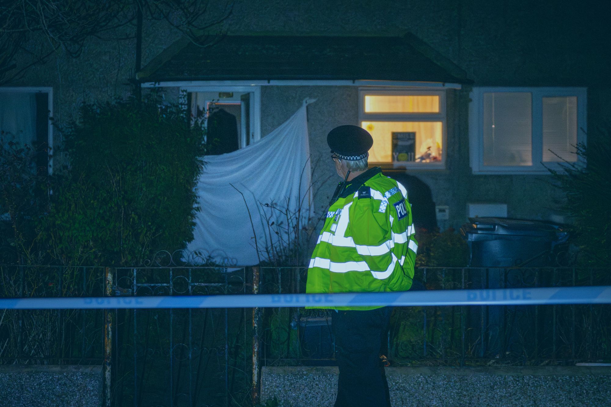 An image collage containing 1 images, Image 1 shows A police officer in a high-visibility vest stands behind police tape in front of a house with a white sheet covering a broken window