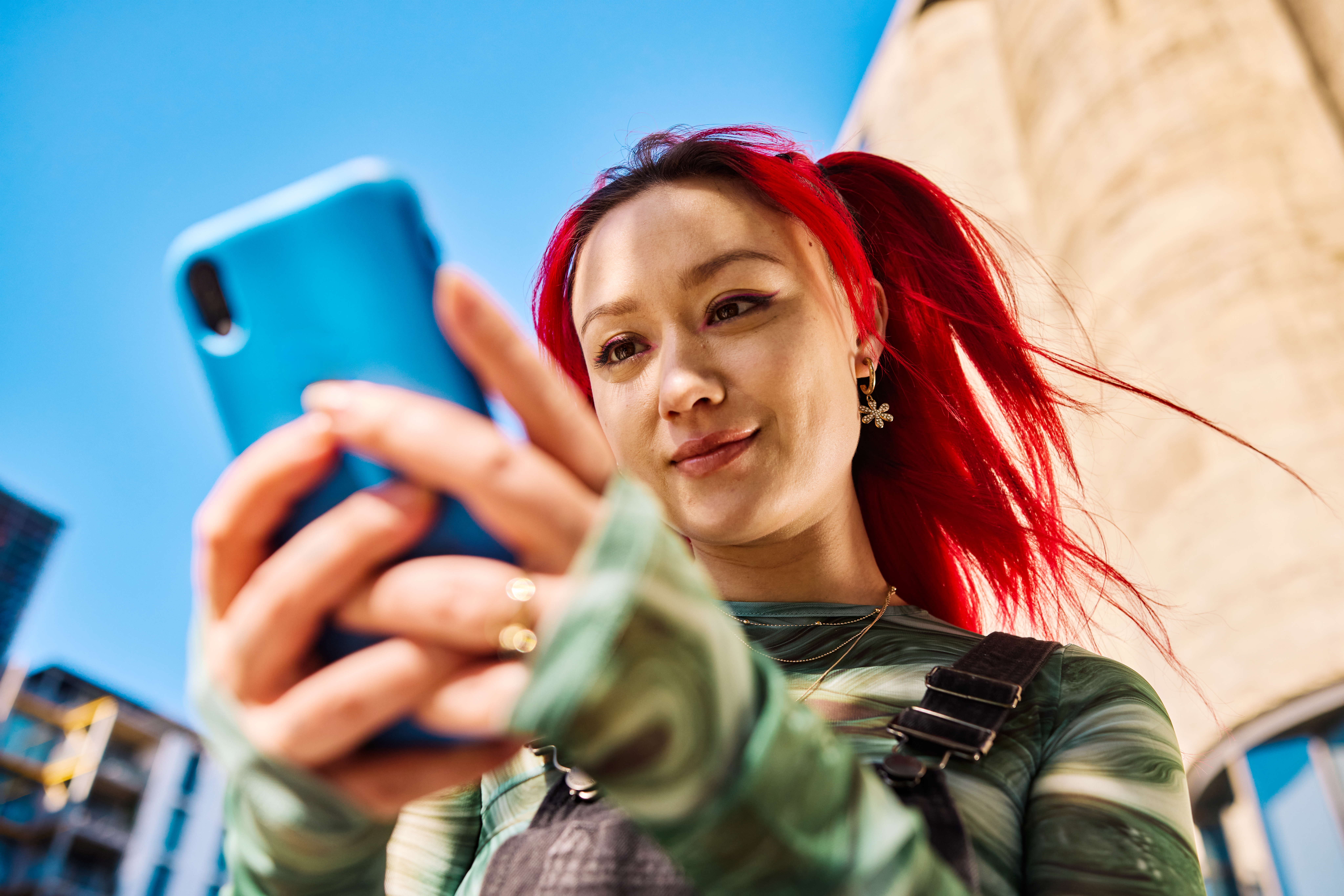 A young woman with bright red hair holds and looks at a smartphone.