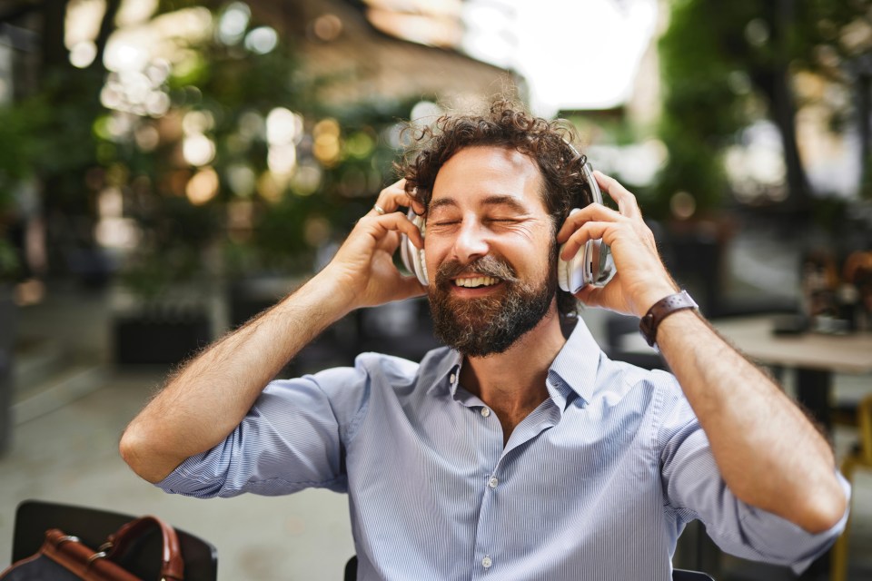 A happy man in an outdoor cafe enjoying music through headphones.