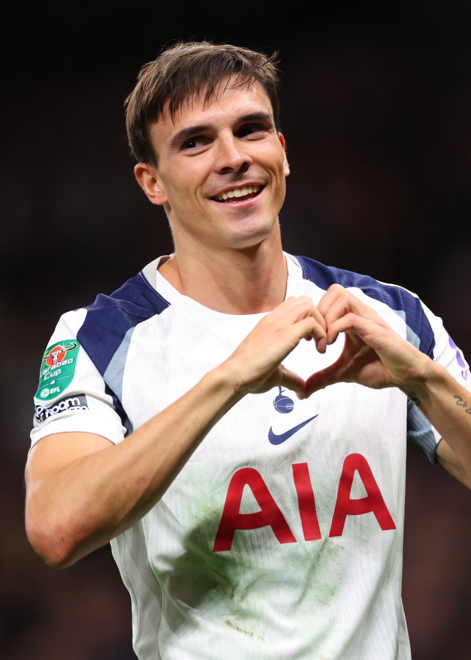 Giovani Lo Celso smiles and forms a heart with his hands while wearing his Tottenham Hotspur uniform.