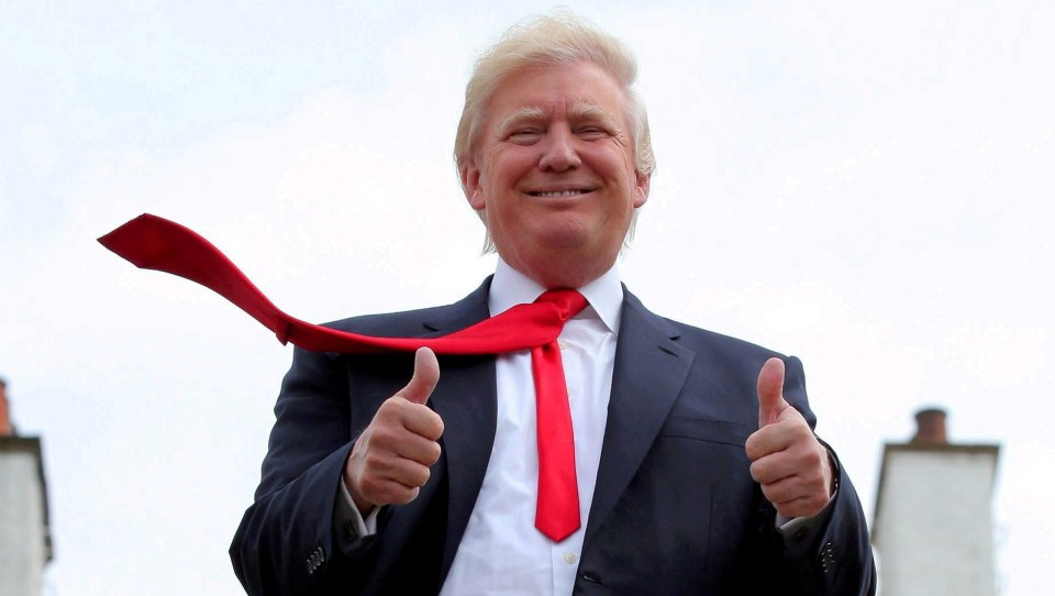 Donald Trump smiles and gives two thumbs up, with his red tie flying over his left shoulder.