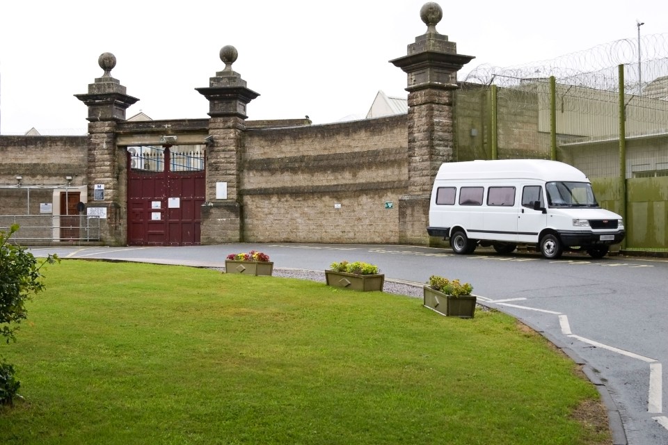 Entrance to HMP Camphill prison in the Isle of Wight, England, showing red gates, stone pillars, a brick wall topped with barbed wire, and a white van.