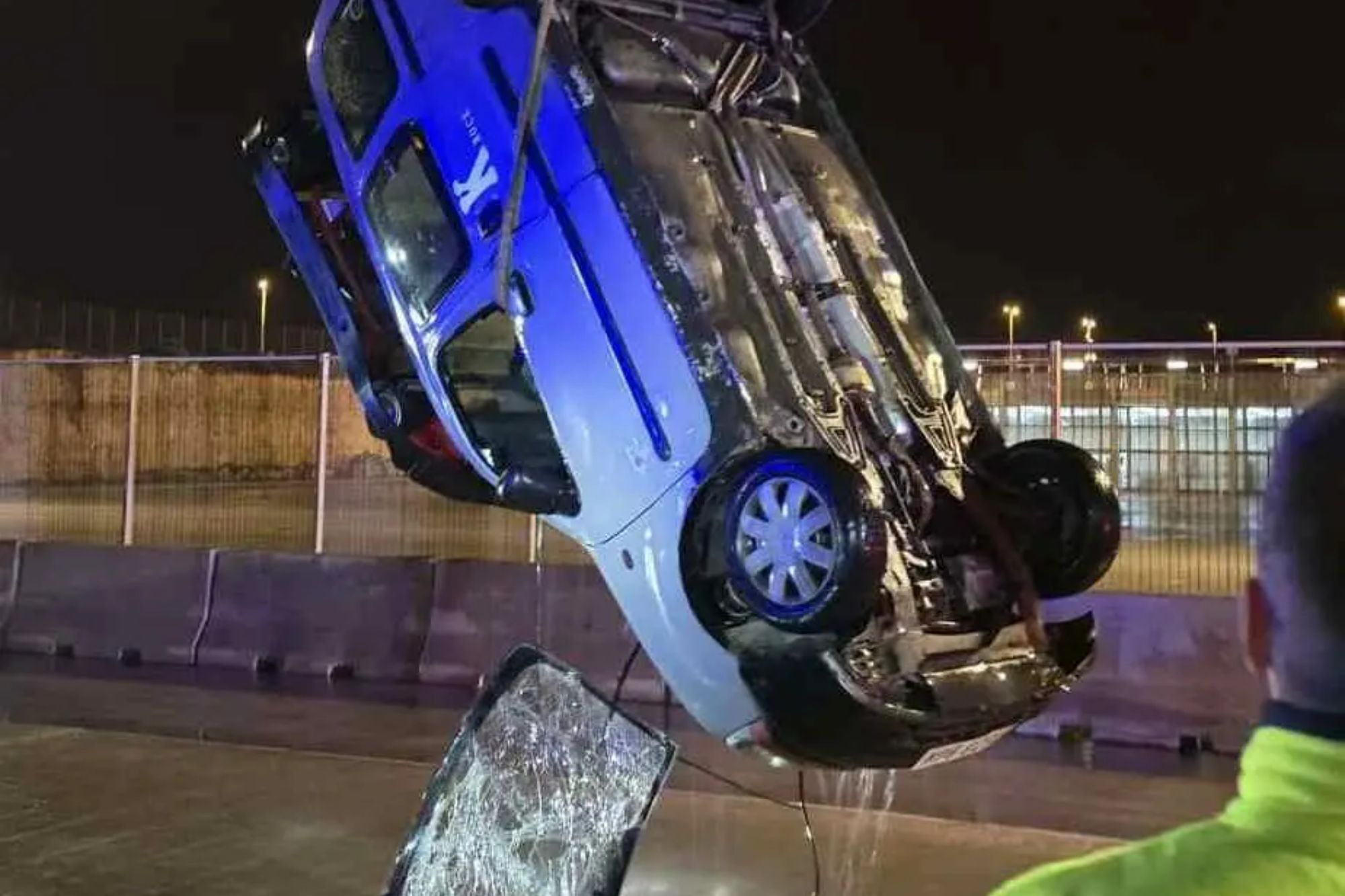 An image collage containing 1 images, Image 1 shows A blue and white car being lifted upside down by a crane from the water at night, with a person in a safety vest watching