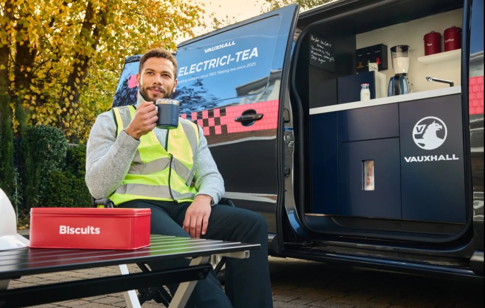 A man in a high-vis vest sitting outside a Vauxhall "Electri-Tea" van, drinking from a mug with a red "Biscuits" tin on the table next to him.