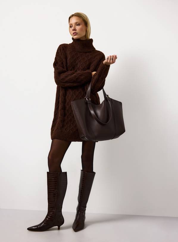 Woman wearing a brown jumper dress, tights, knee-high crocodile boots, and holding a brown tote bag.